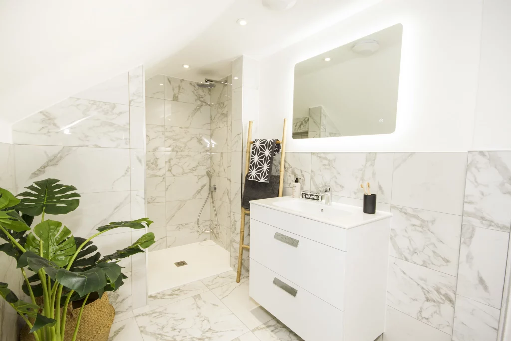 Bright white marble-tiled bathroom with walk-in shower, floating sink cabinet, illuminated mirror, ladder towel rack and potted green plant