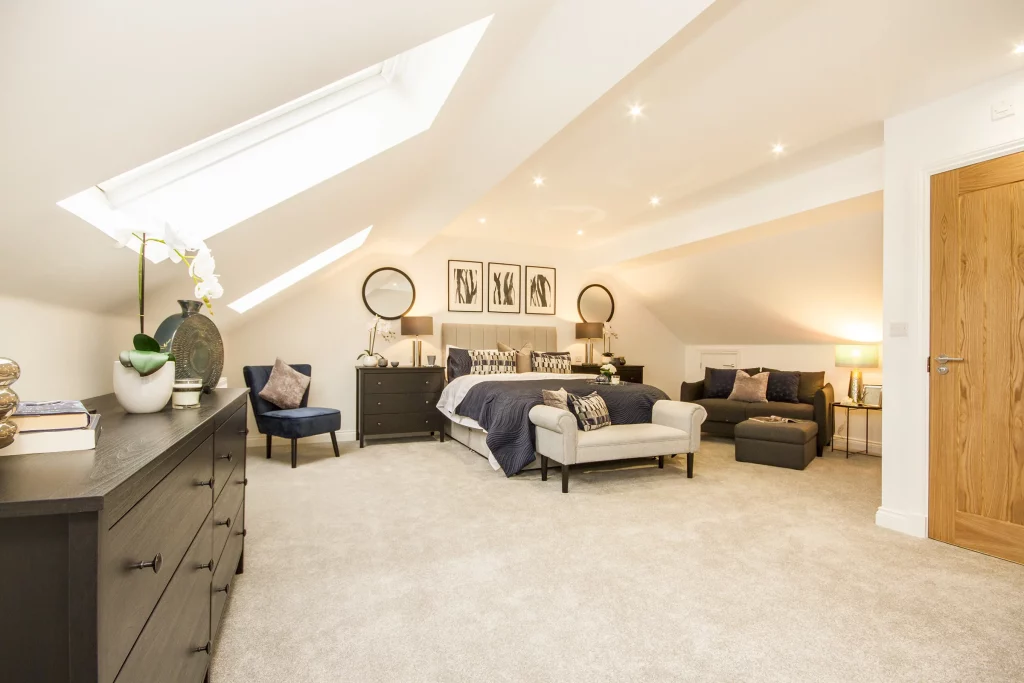 Spacious, softly lit attic bedroom with sloped skylights, king bed with navy throw, armchair, dresser and cozy seating area.