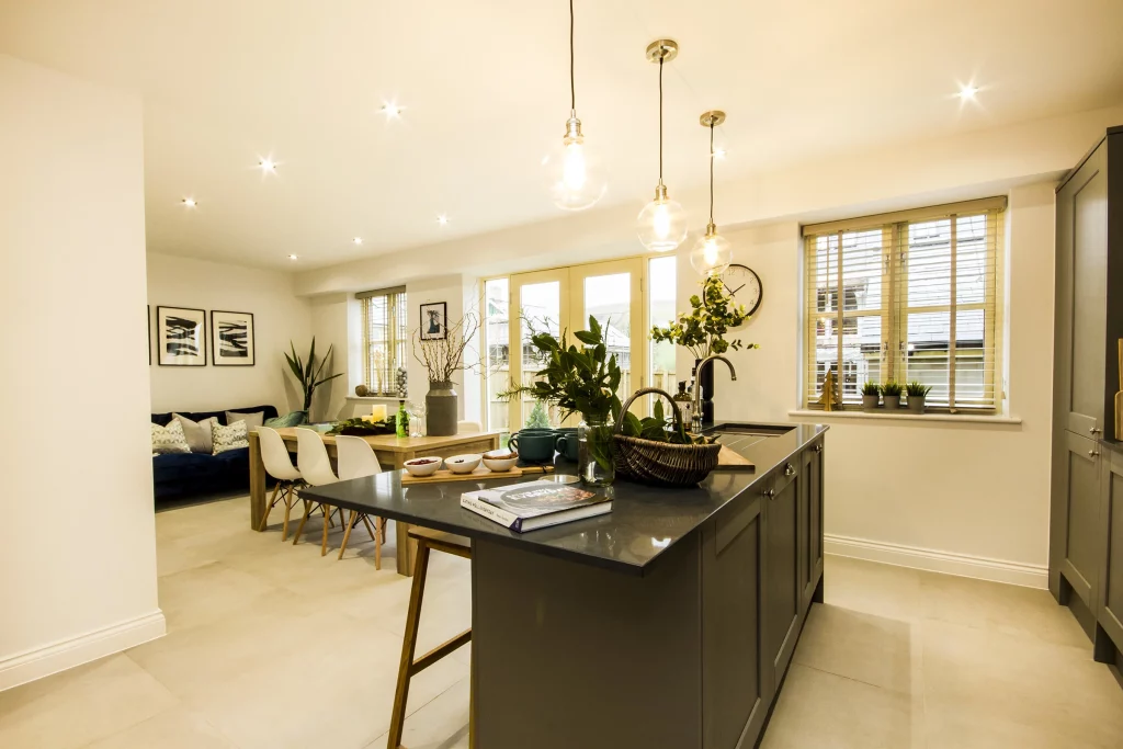 Open-plan kitchen and dining area with a dark island countertop, hanging globe pendant lights, dining table with white chairs and indoor plants.