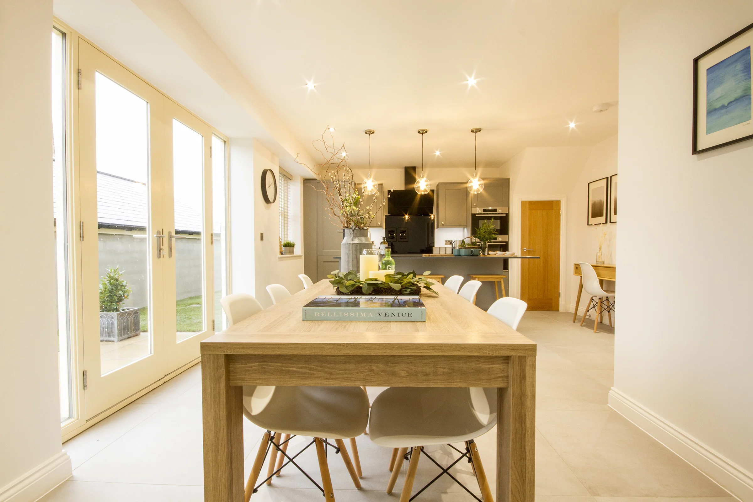 Long wooden dining table with white chairs, a plant centerpiece and books, leading to a modern open kitchen with pendant lights.