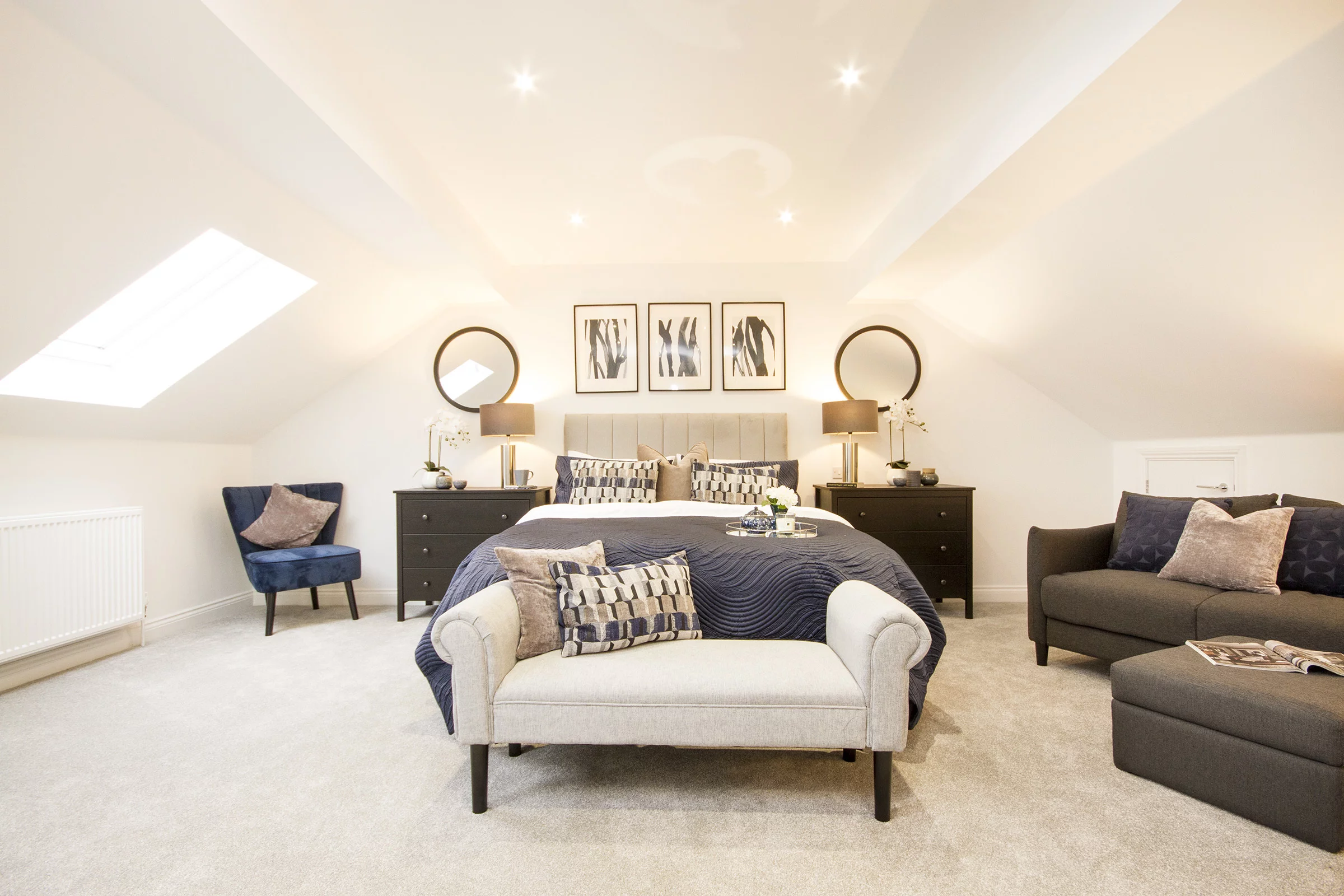 Spacious, neutral-toned attic bedroom with a central bed, bench, navy quilt, pair of nightstands with lamps and round mirrors
