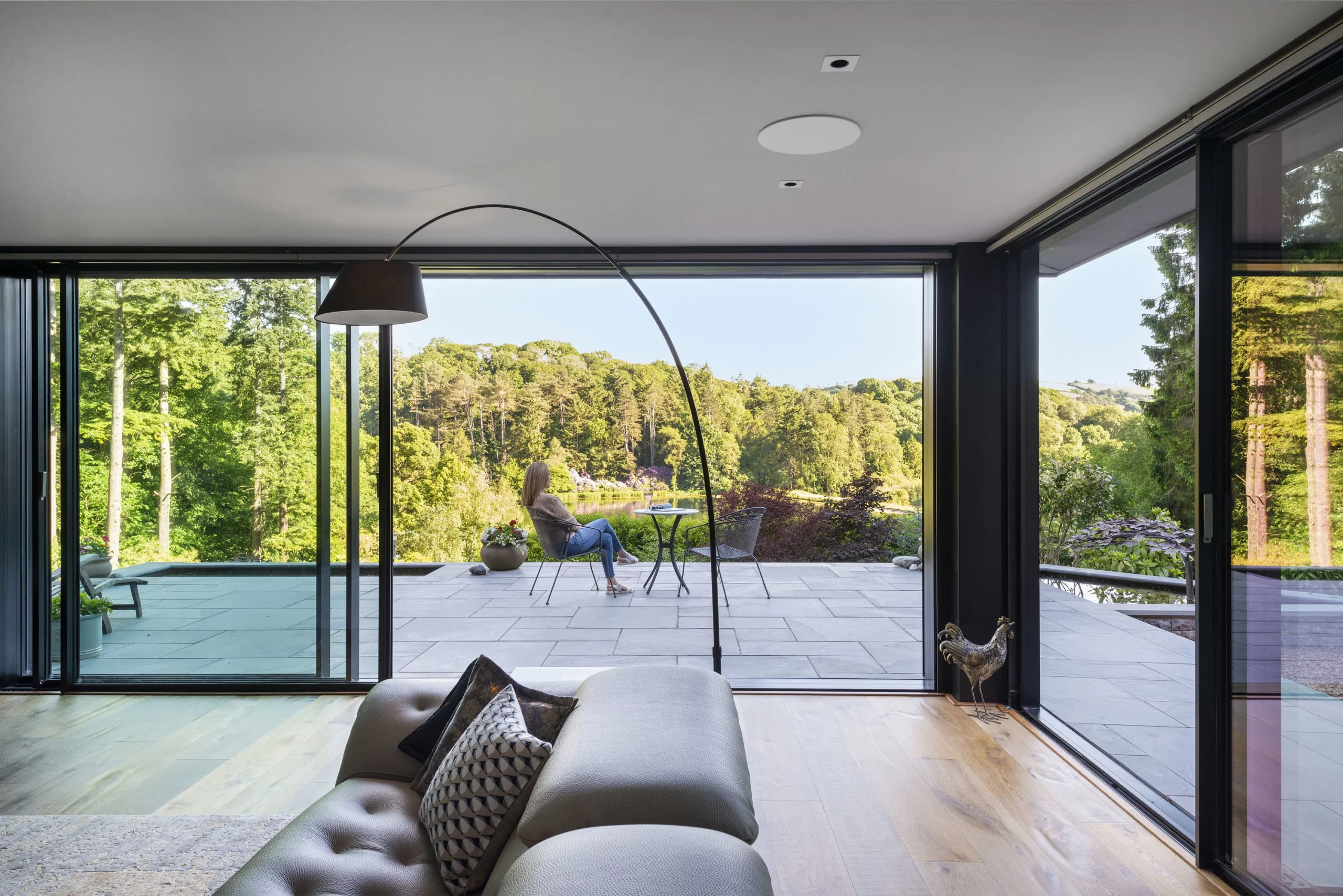 Modern living room looking through wide glass doors to a terrace where a woman sits at a small table overlooking wooded hills