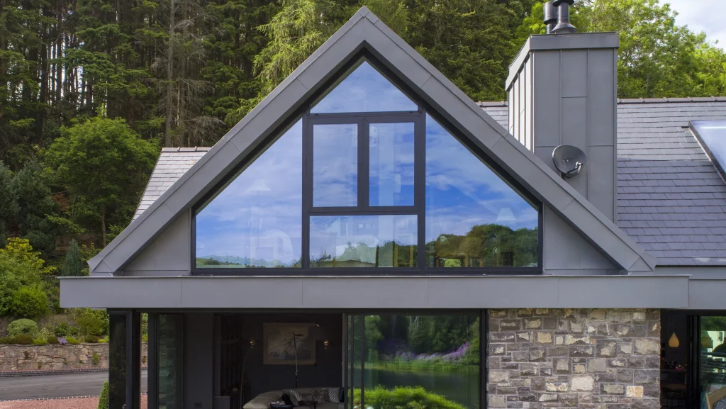 Modern gabled house facade with large triangular glass window reflecting sky and trees, grey cladding and stone wall below.