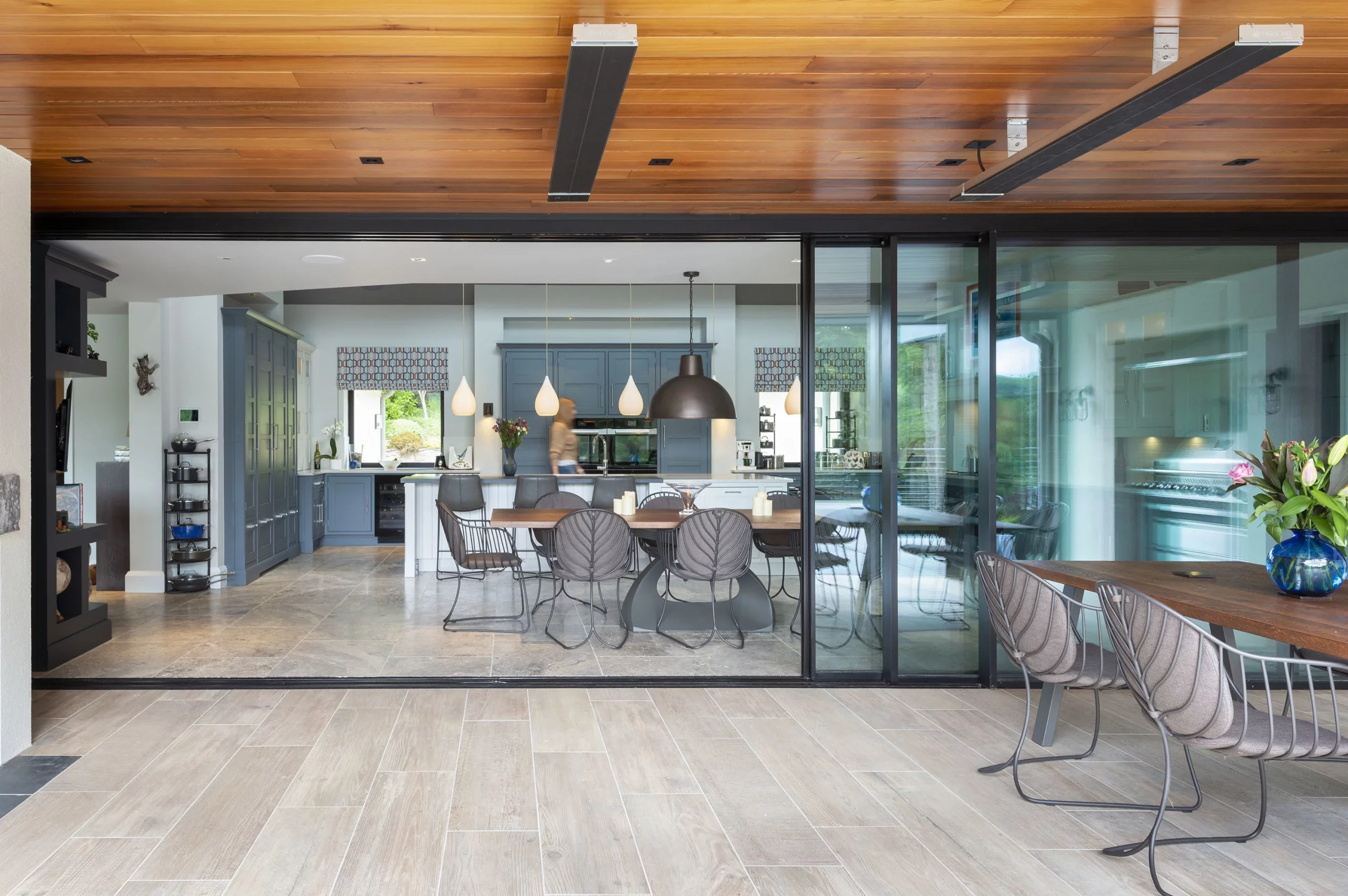 Open-plan dining and kitchen seen through large glass sliding doors, wooden ceiling, modern pendant lights and grey chairs
