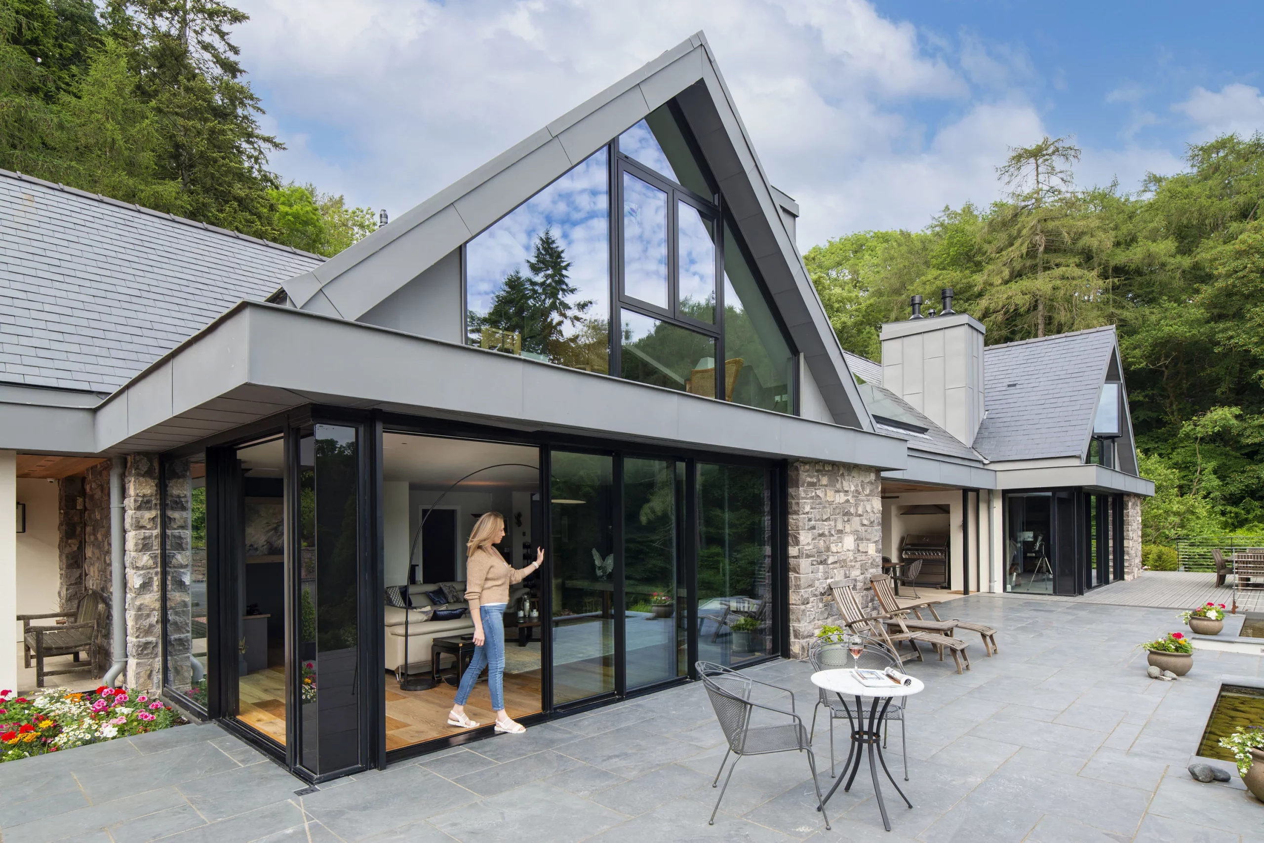 Modern A-frame house with large glass windows opening onto a patio where a woman steps outside among chairs and planters