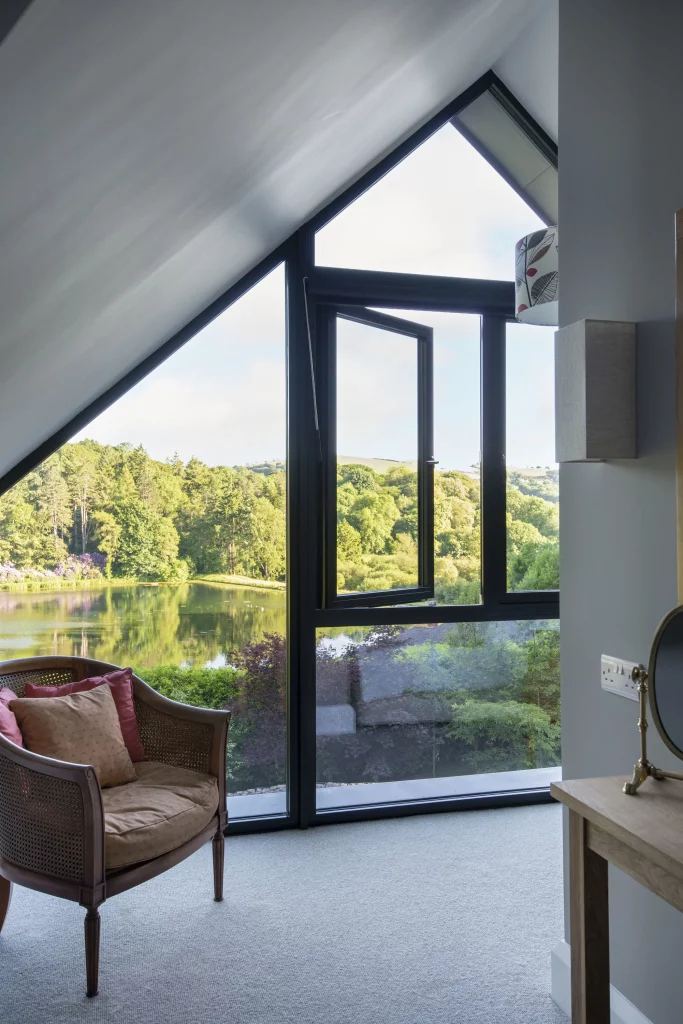 Corner room with a large triangular window overlooking a lake and green trees, a cushioned wicker armchair by the glass.