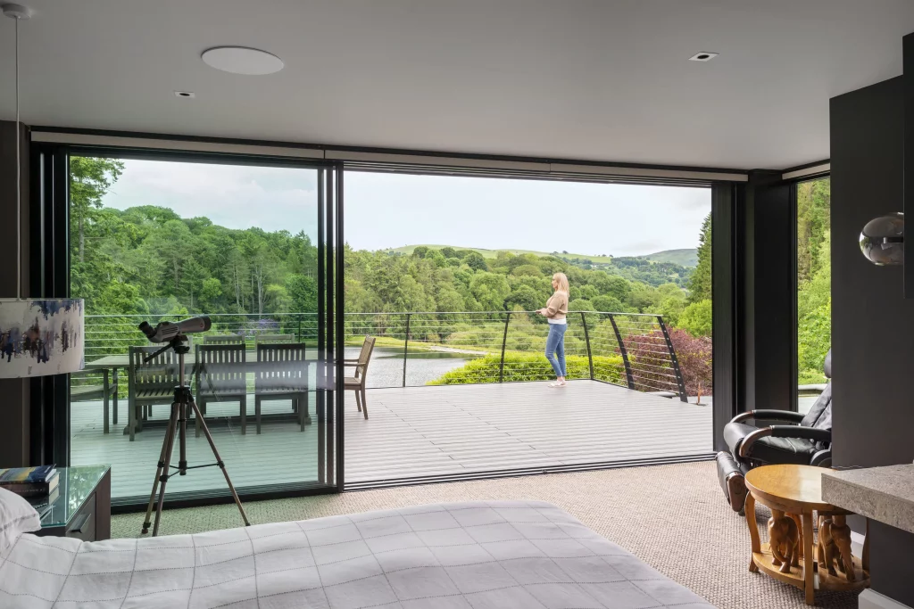 Bedroom interior with open sliding glass doors onto a deck where a woman stands looking over a green lakeside valley.