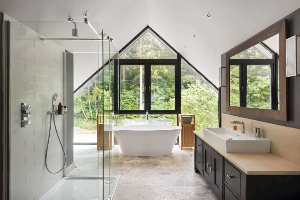 Light, modern bathroom with freestanding tub under triangular window, glass shower on left and sink with wooden mirror on right