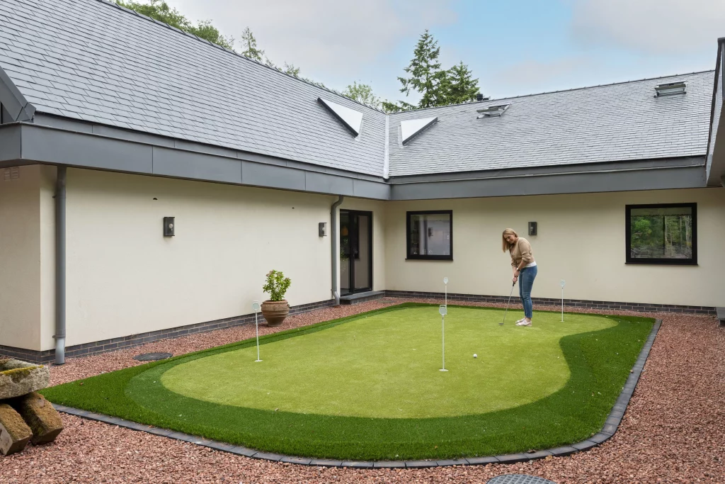 Woman practising putting on a small backyard artificial grass green surrounded by cream house walls and pebble border