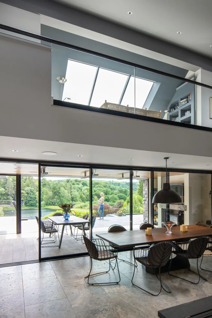 Modern open-plan dining room with large glass doors opening to terrace and green lake view, balcony and loft above