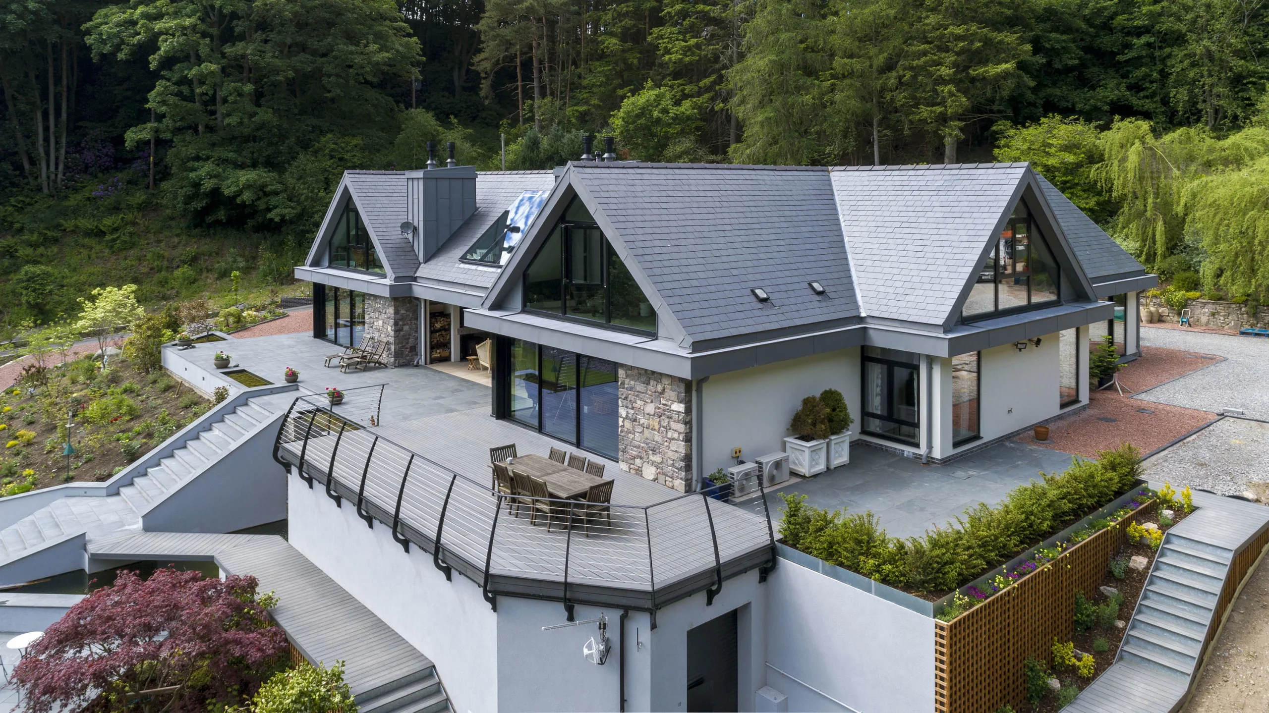 Modern slate-roofed house with large triangular glass gables, wraparound terrace and outdoor dining area set against a wooded hillside