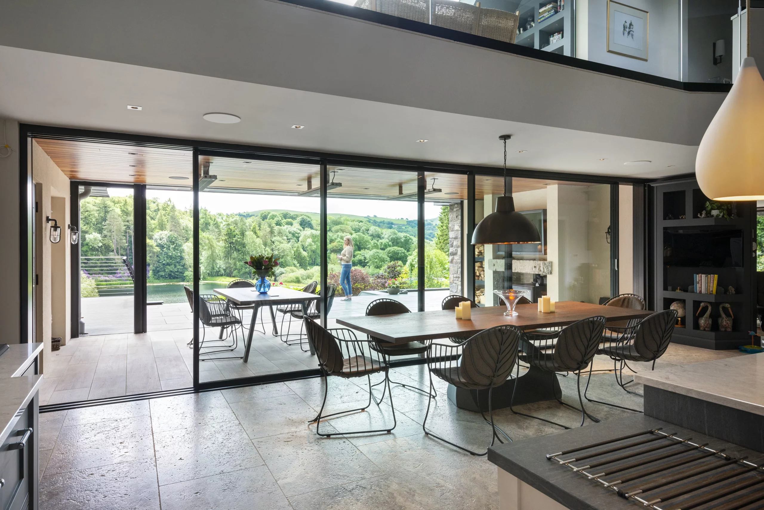Spacious modern dining area with long wooden table, black pendant lights and glass sliding doors opening to a terrace and green hillside.