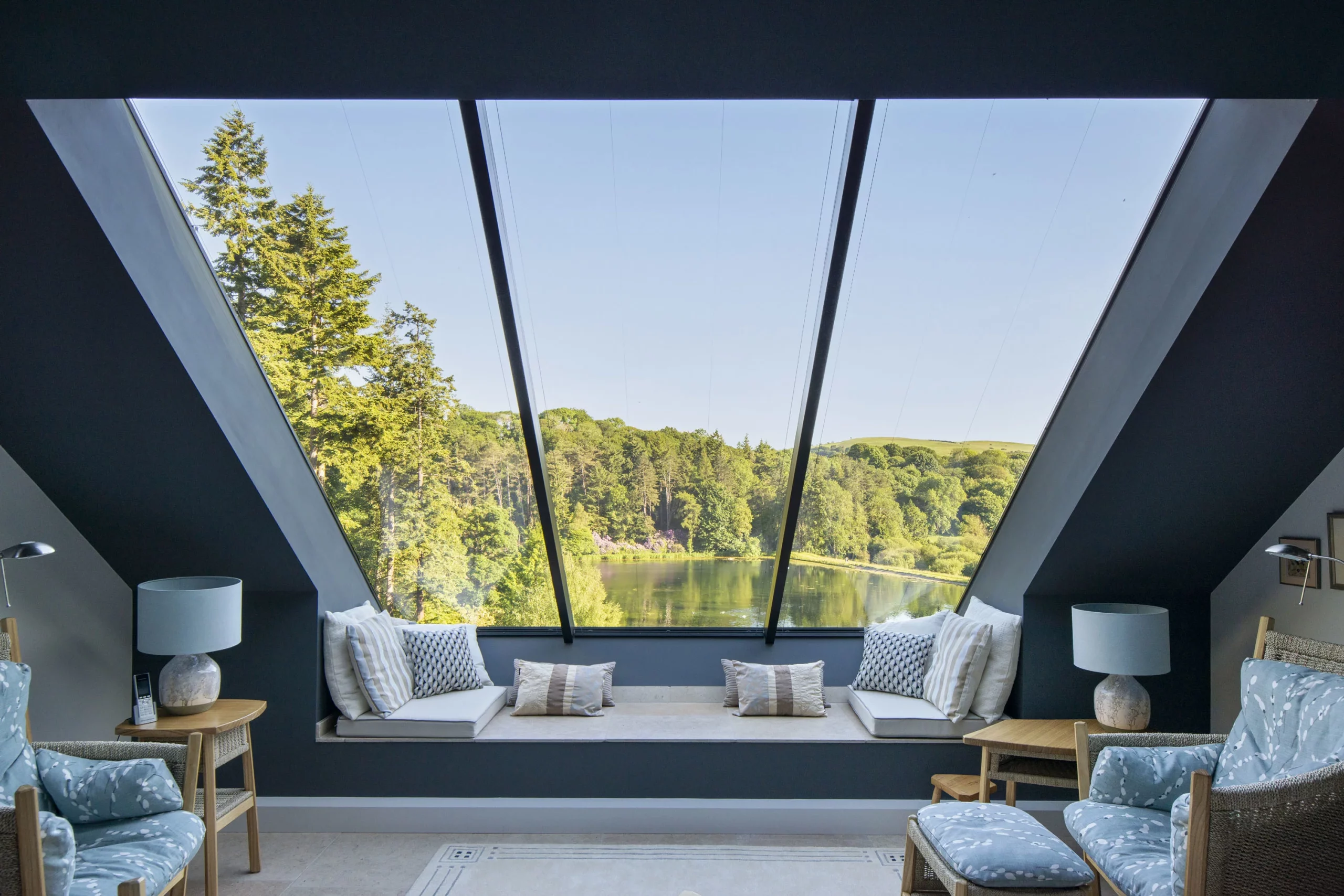 Cozy attic seating area with cushions facing a large triangular skylight framing trees and a lake view.