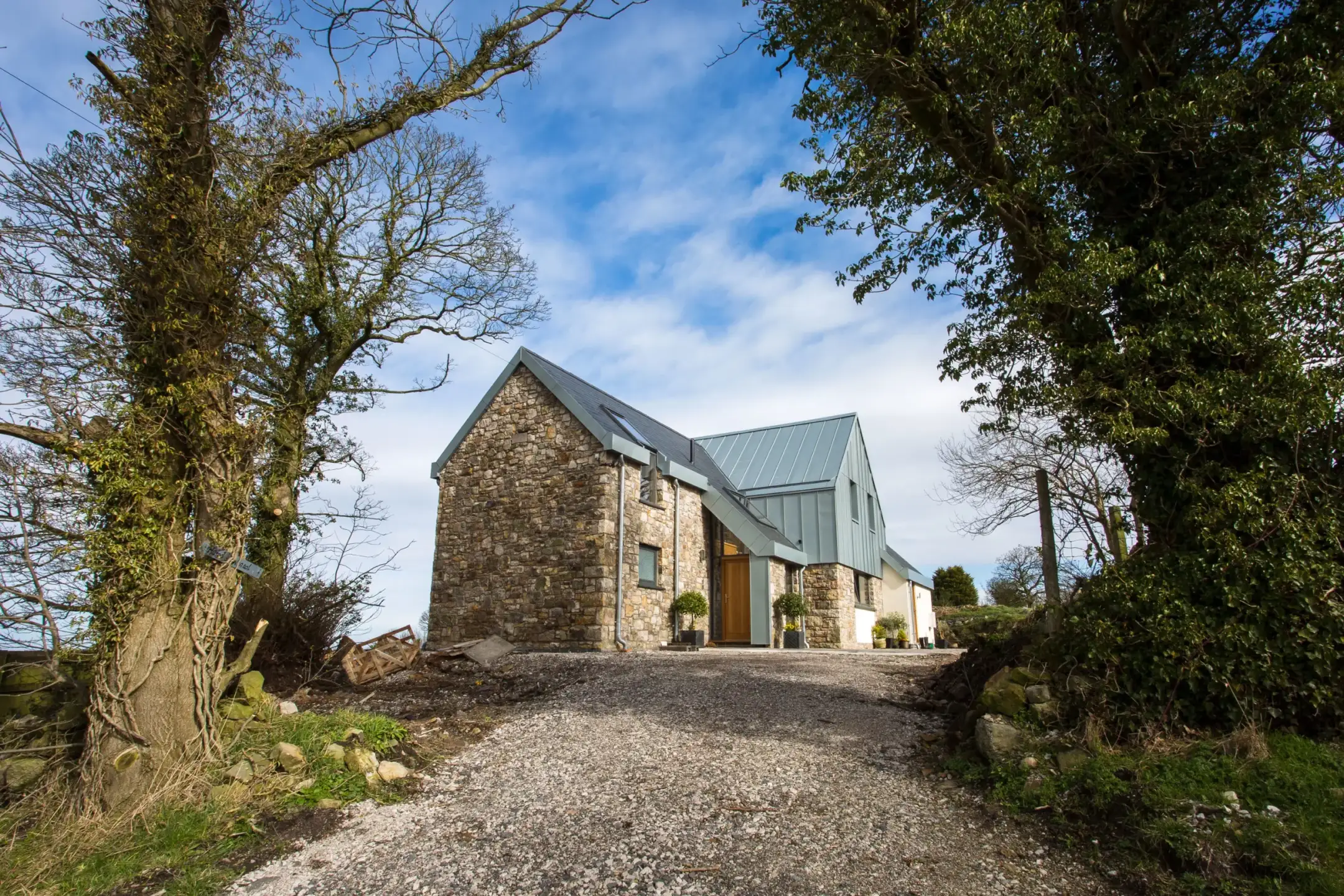 Stone cottage with modern metal roof and wooden door at the end of a gravel drive, framed by large trees and blue sky