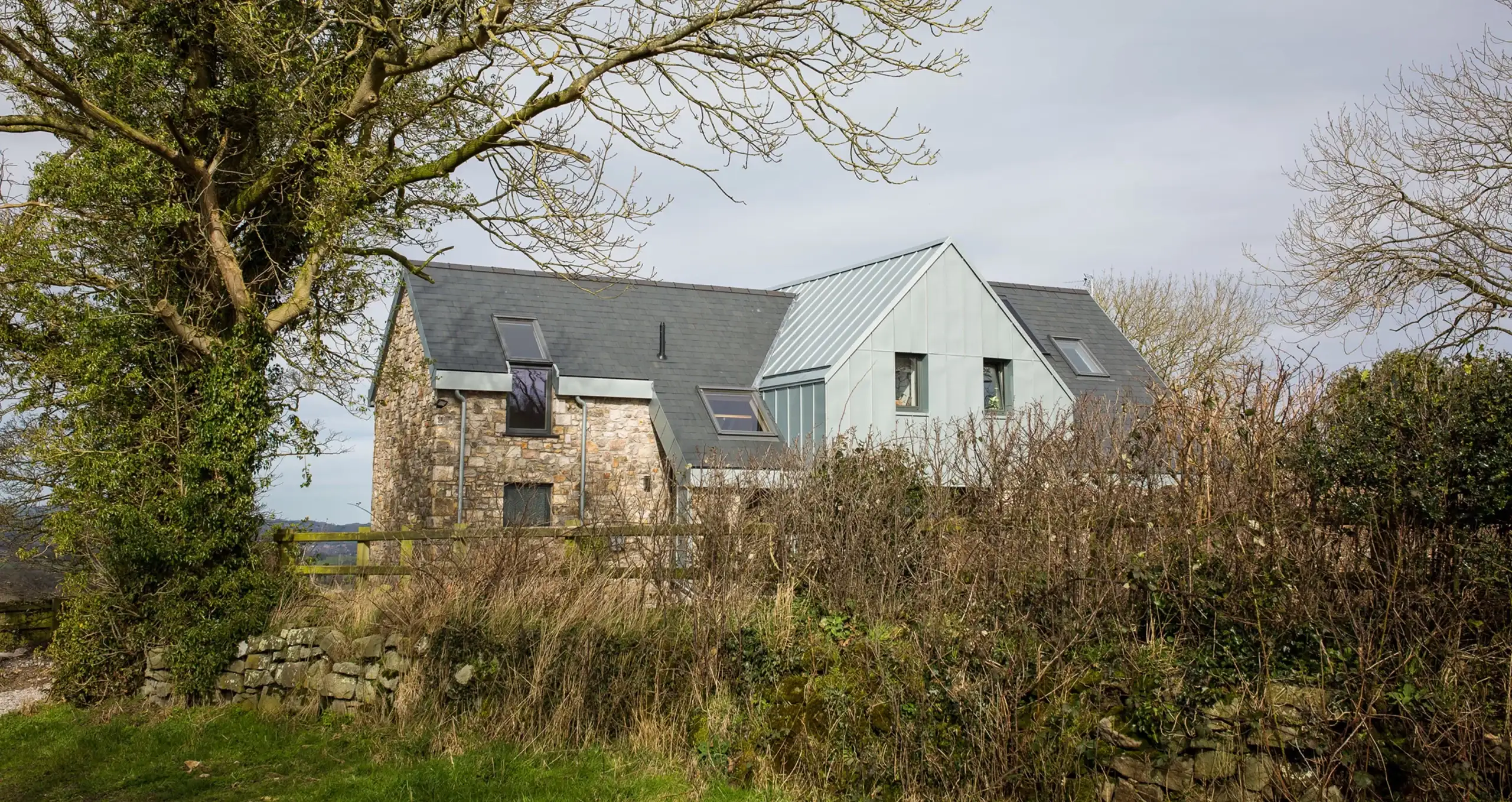 Stone and metal-clad rural house with slate roofs and dormer windows behind a hedgerow, framed by large bare trees.