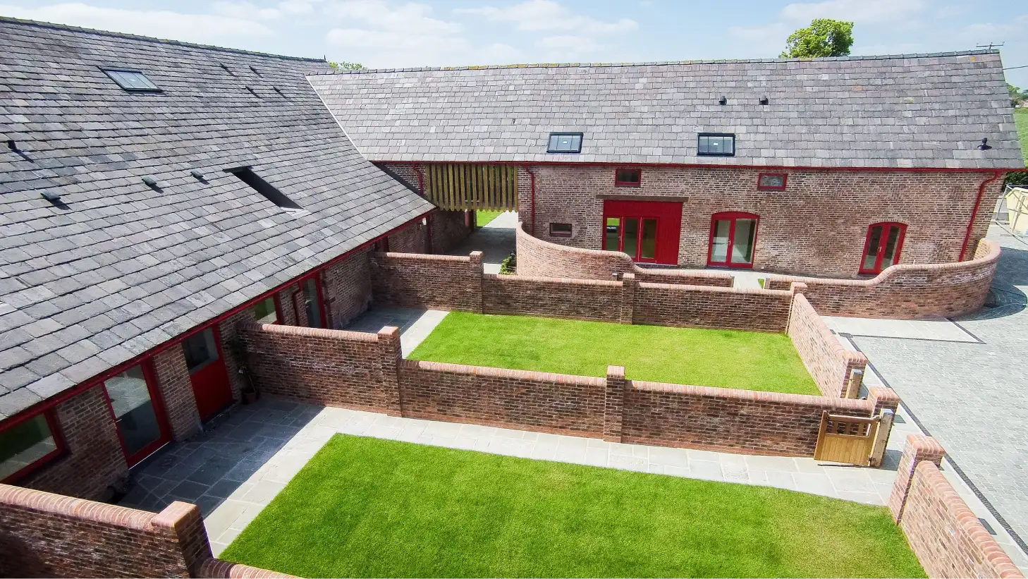 Courtyard of converted red-brick barns with slate roofs, red doors and windows, paved paths and small enclosed lawns.