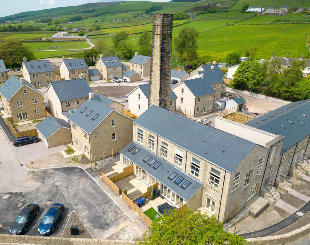 New stone-built houses with grey roofs clustered around a tall old brick chimney, with green fields and hills in the background.