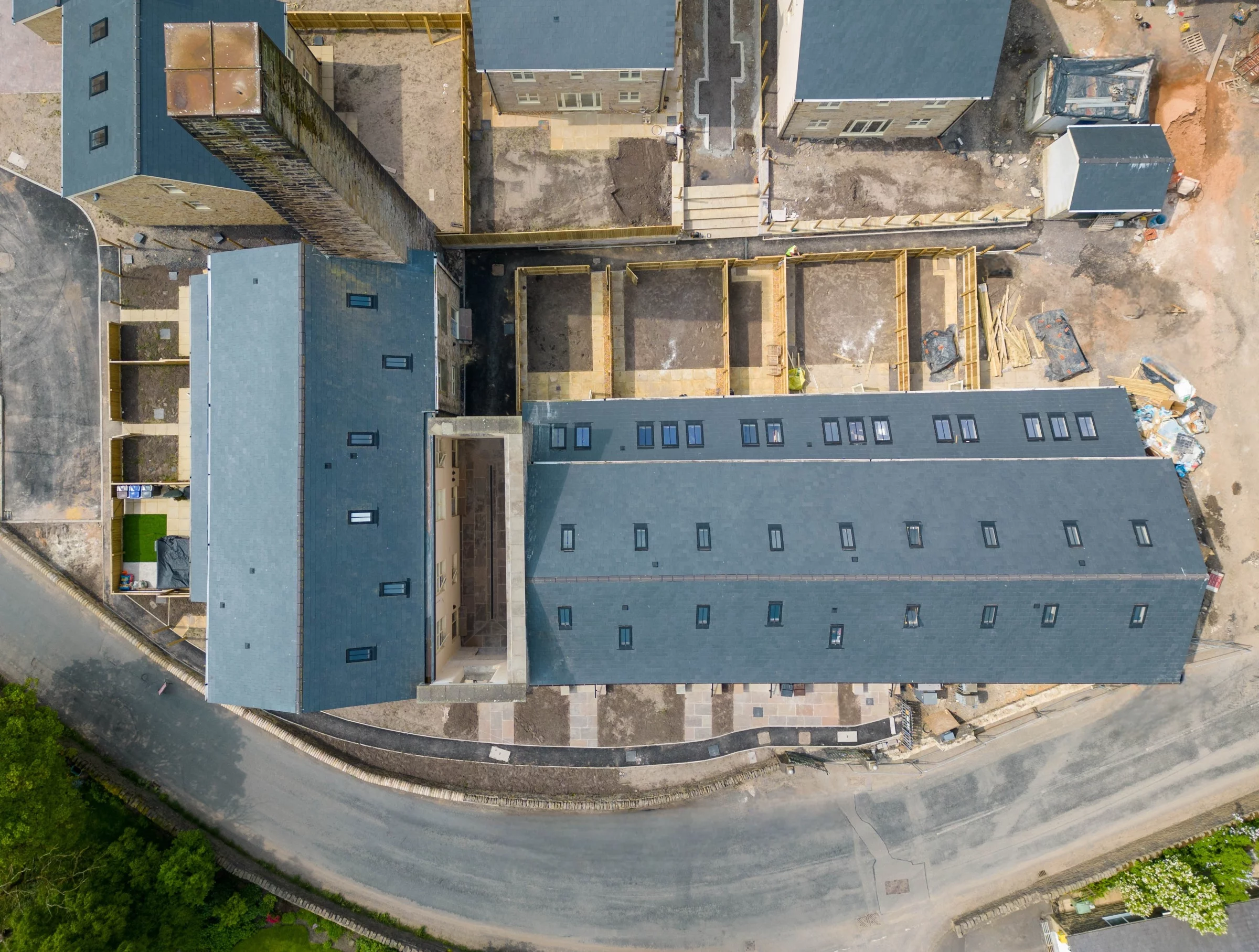 Aerial view of a residential construction site showing rooftop rows, courtyards and fenced foundations next to a curved road
