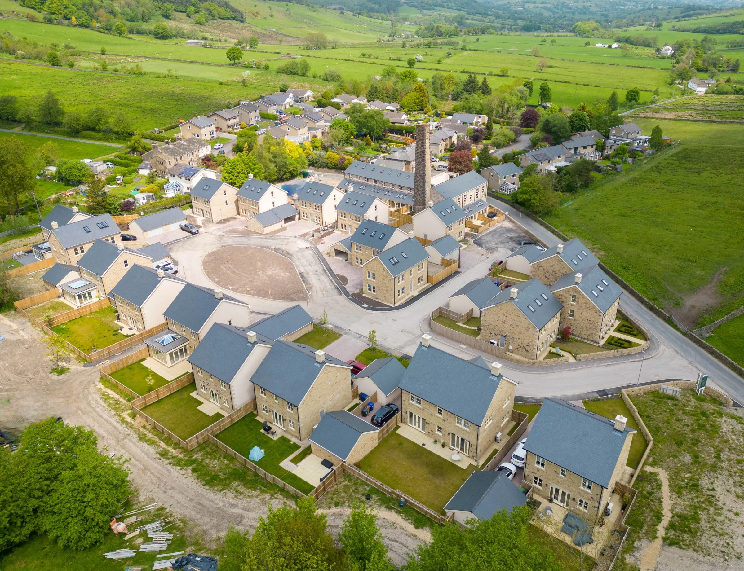 Aerial view of a new housing development with stone houses, grey roofs, fenced backyards and surrounding green fields.