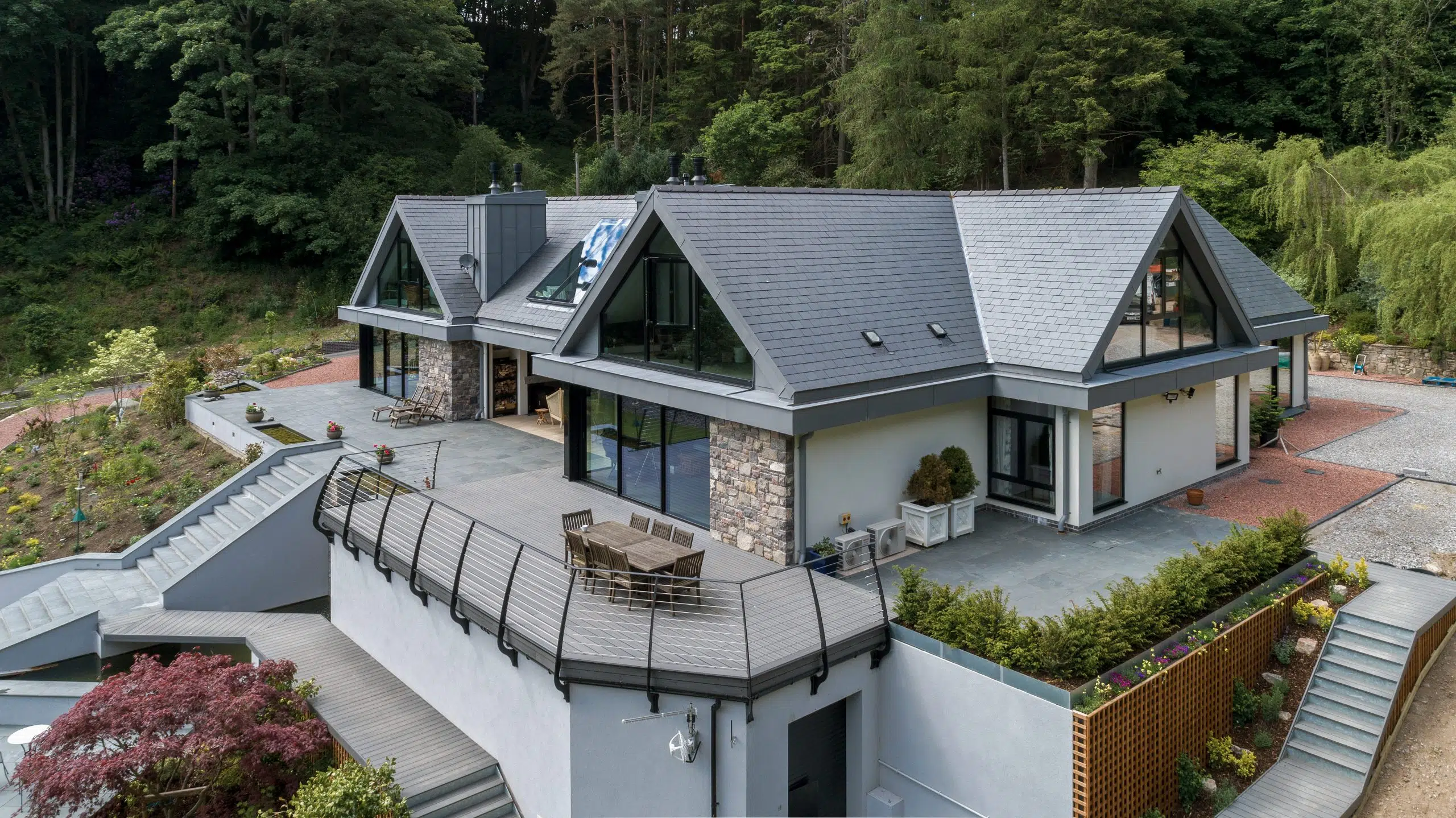 Modern hillside house with slate gabled roofs, large glass windows and a terrace with outdoor dining table.