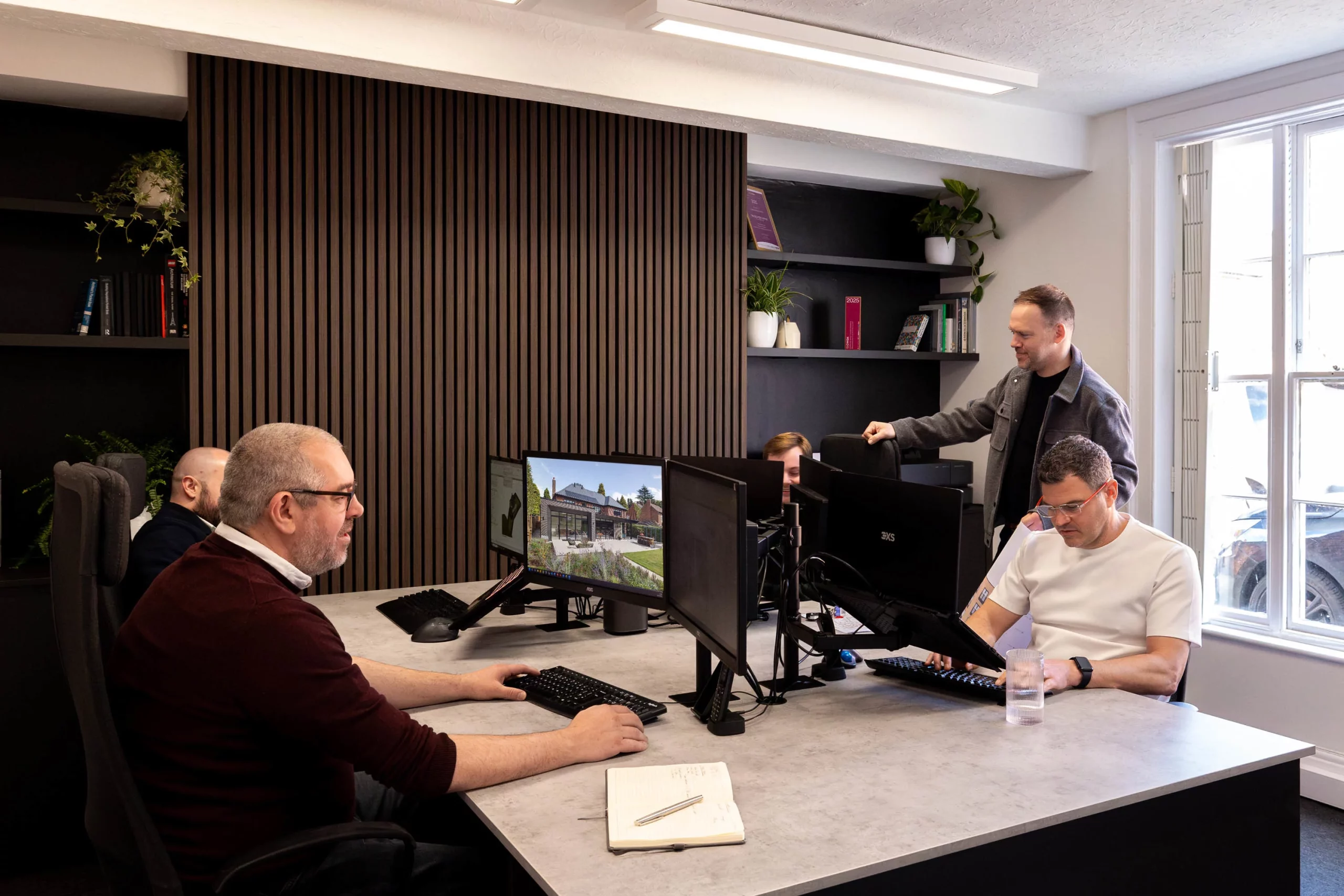 Office scene with five colleagues working at desks and computers, wood-paneled wall and shelves with plants and books in the background.
