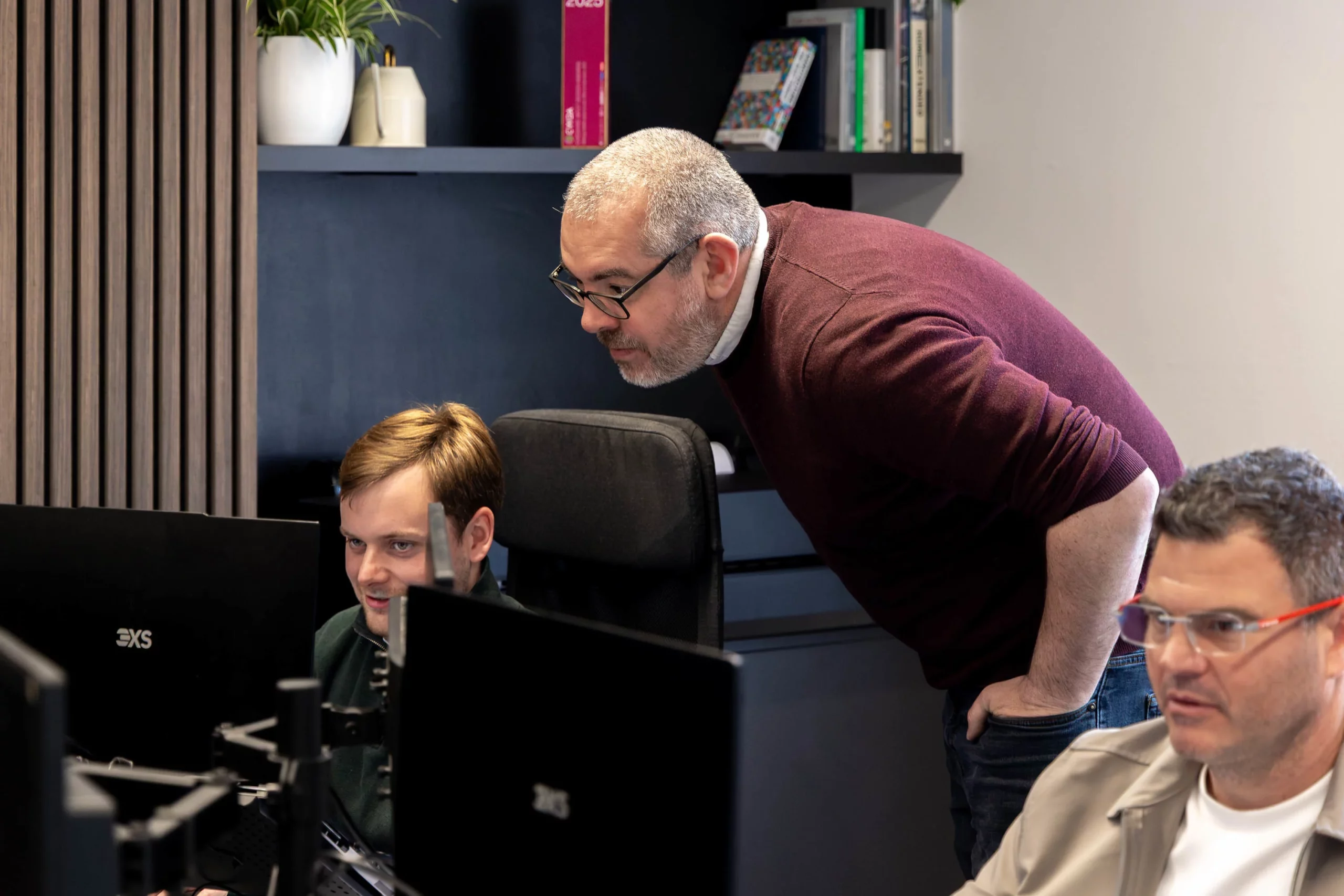 Man in glasses and maroon sweater leaning over a seated colleague's monitor while two other coworkers work at nearby screens in an office.