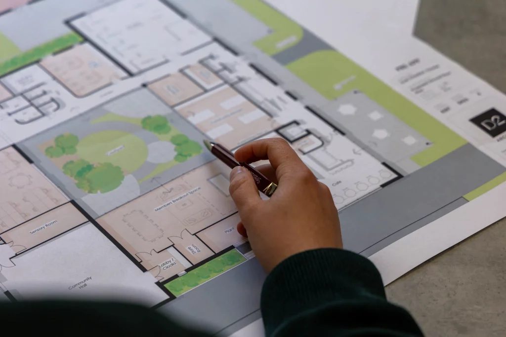 Hand holding a pen pointing at a colour architectural floor plan with labelled rooms and green courtyard areas.