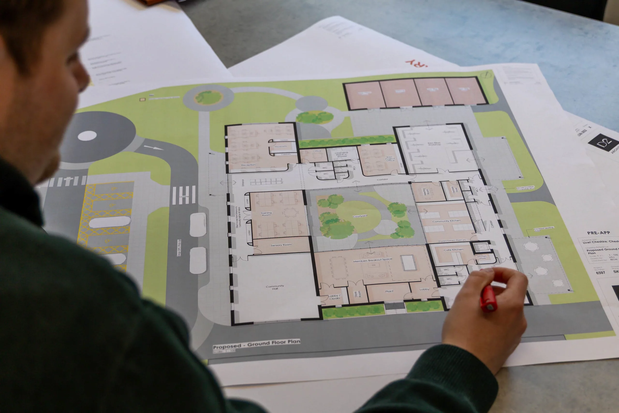 Person holding a red pen reviewing a large printed architectural ground-floor plan with rooms, courtyard and parking layout.