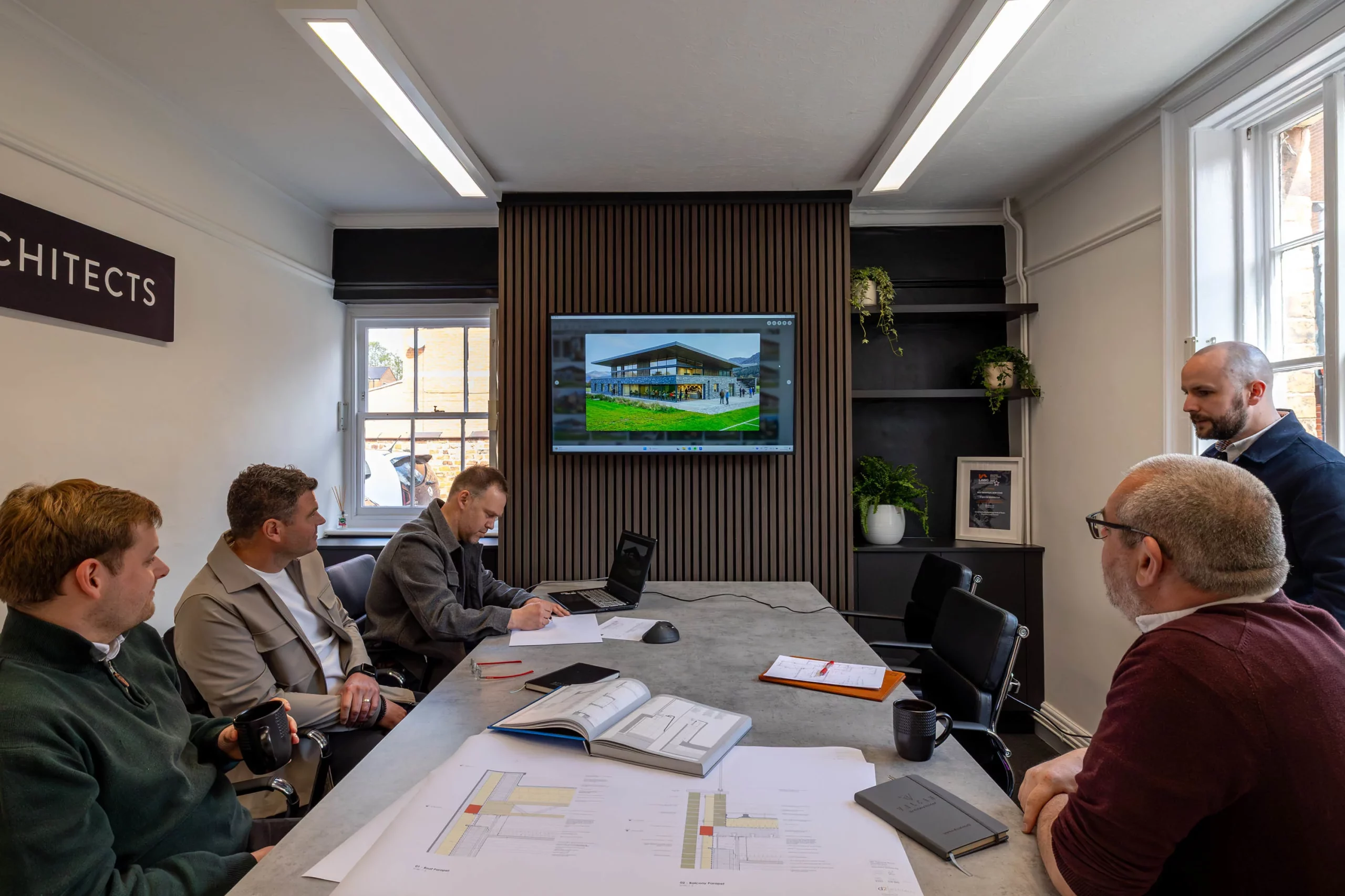 Five architects around a conference table studying plans and a laptop while a screen on a wood-slatted wall displays a building design.