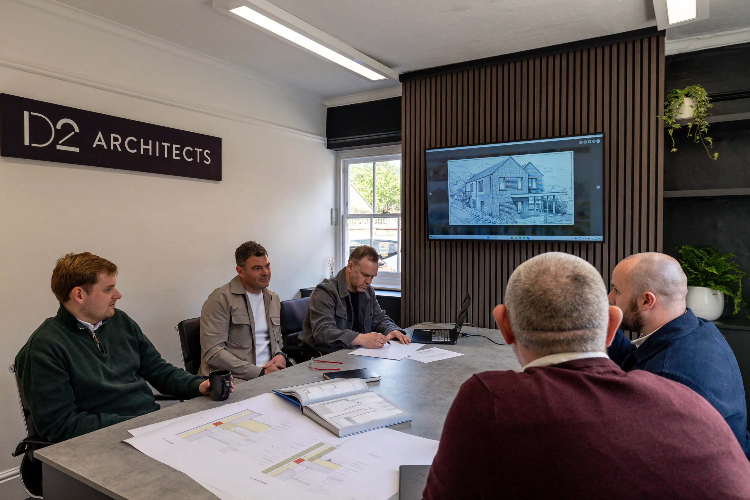 Five men around a meeting table reviewing architectural plans, screen on wall shows a house drawing and a sign reads "D2 Architects".