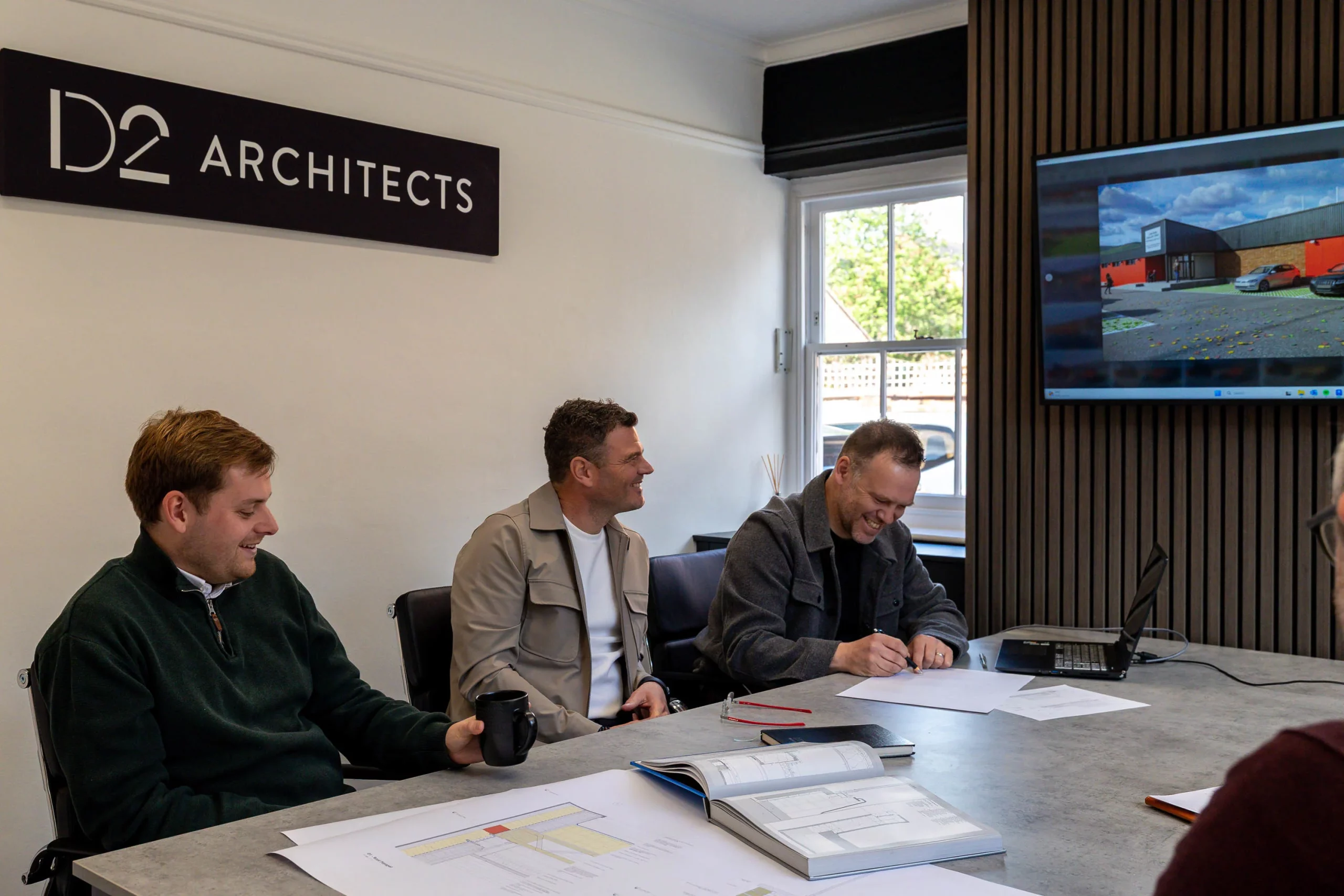 Three men in an office meeting smiling and signing papers beneath a wall sign reading D2 Architects, plans on the table.