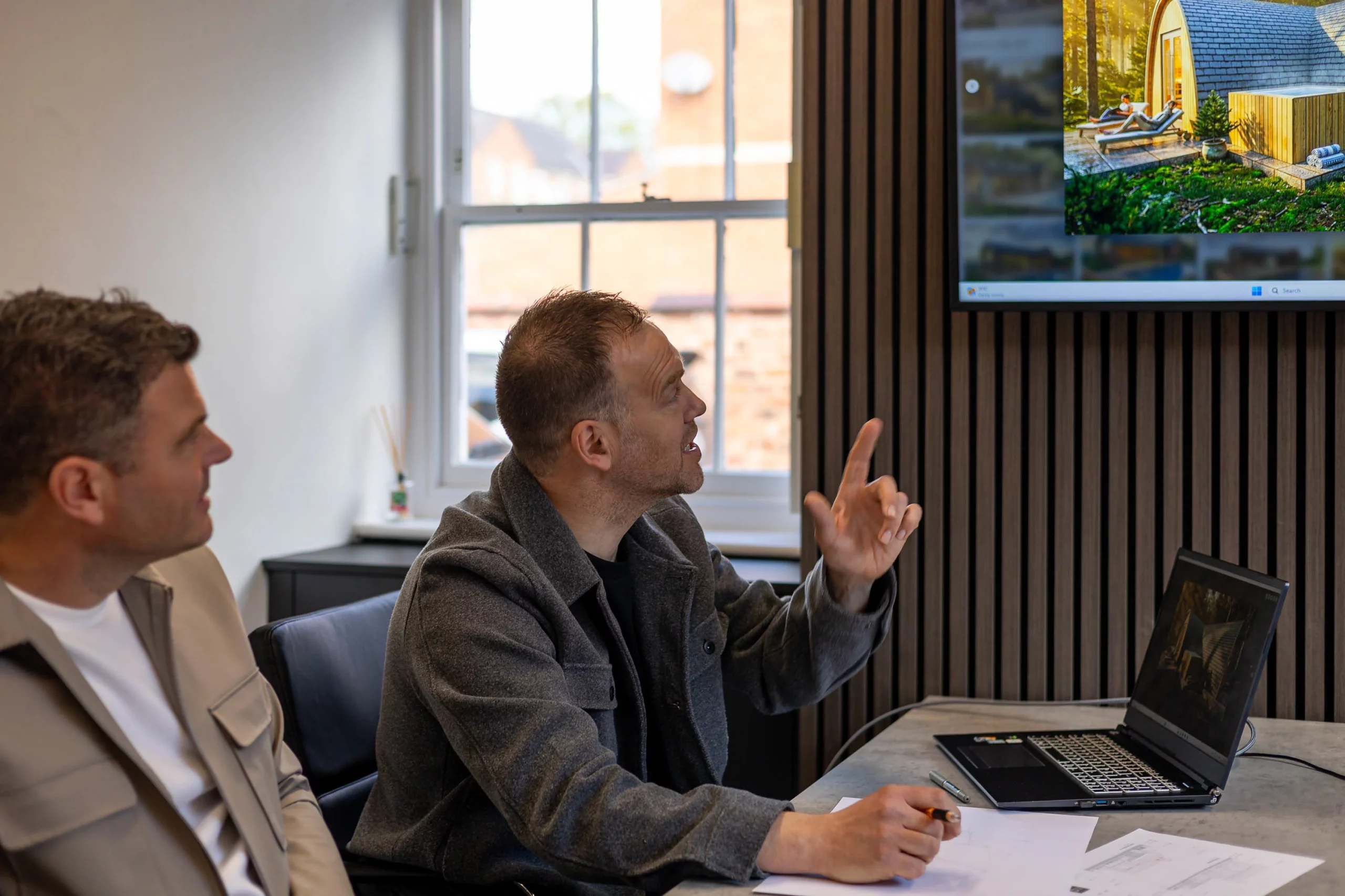 Two men in a meeting room looking at a wall-mounted screen, one pointing up while a laptop and papers sit on the table.