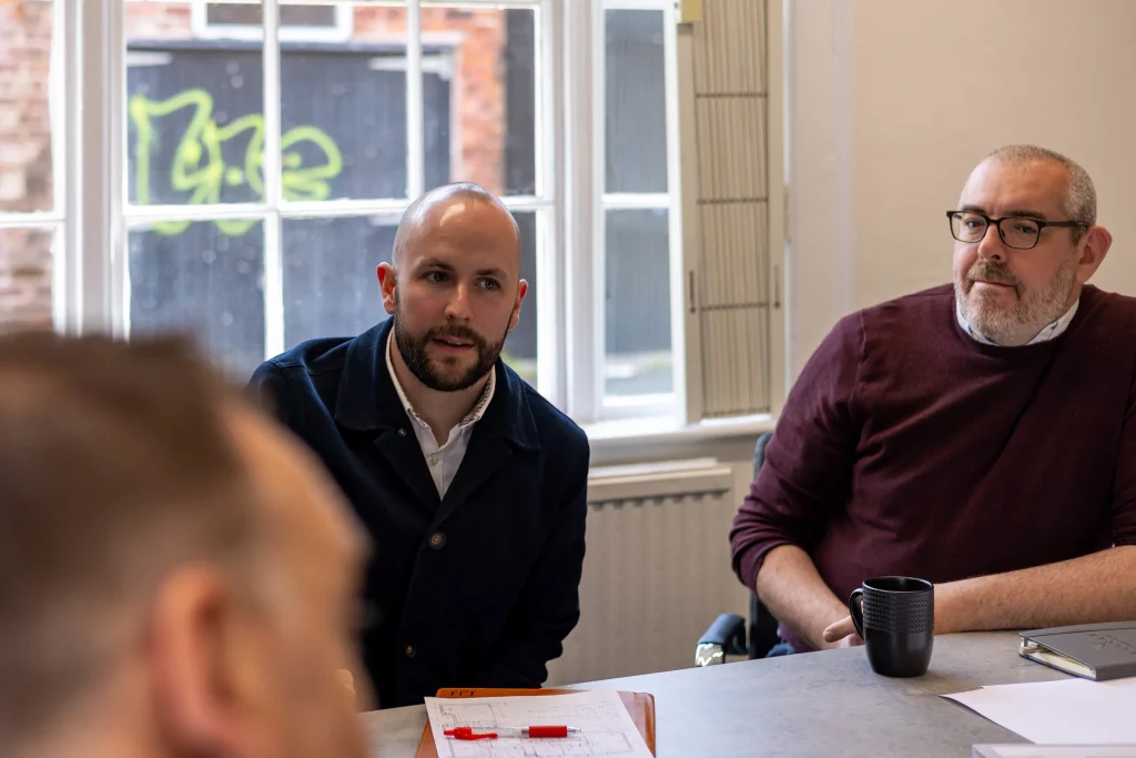 Two men seated at a meeting table near a window, one bald with a beard listening attentively and the other wearing glasses holding a black mug.