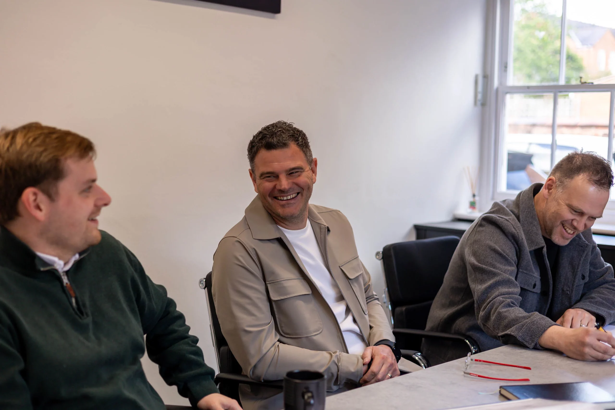 Three men sitting at a table in a bright meeting room, smiling and laughing during a casual discussion.