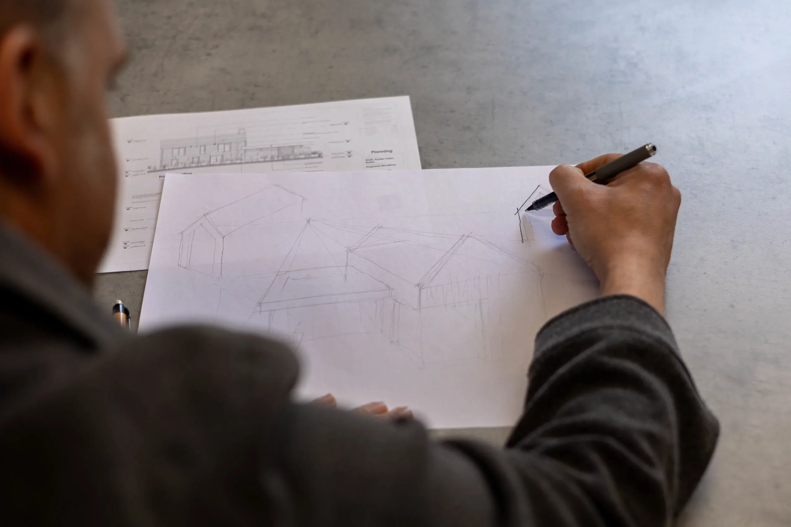 Person sketching roofed building forms on paper with a pen, architectural plan visible underneath on the table.