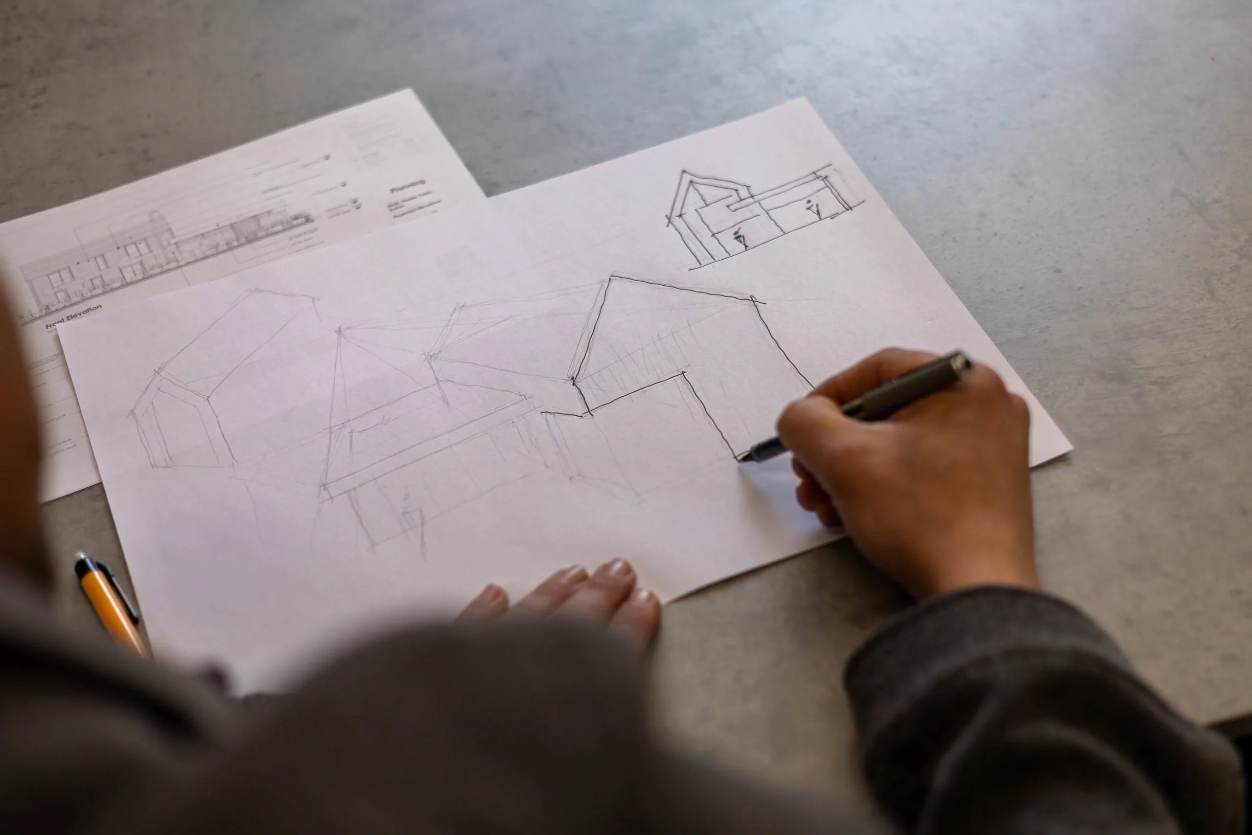 Hand sketch of a house on paper with a hand holding a pen, faint architectural plans visible beneath.