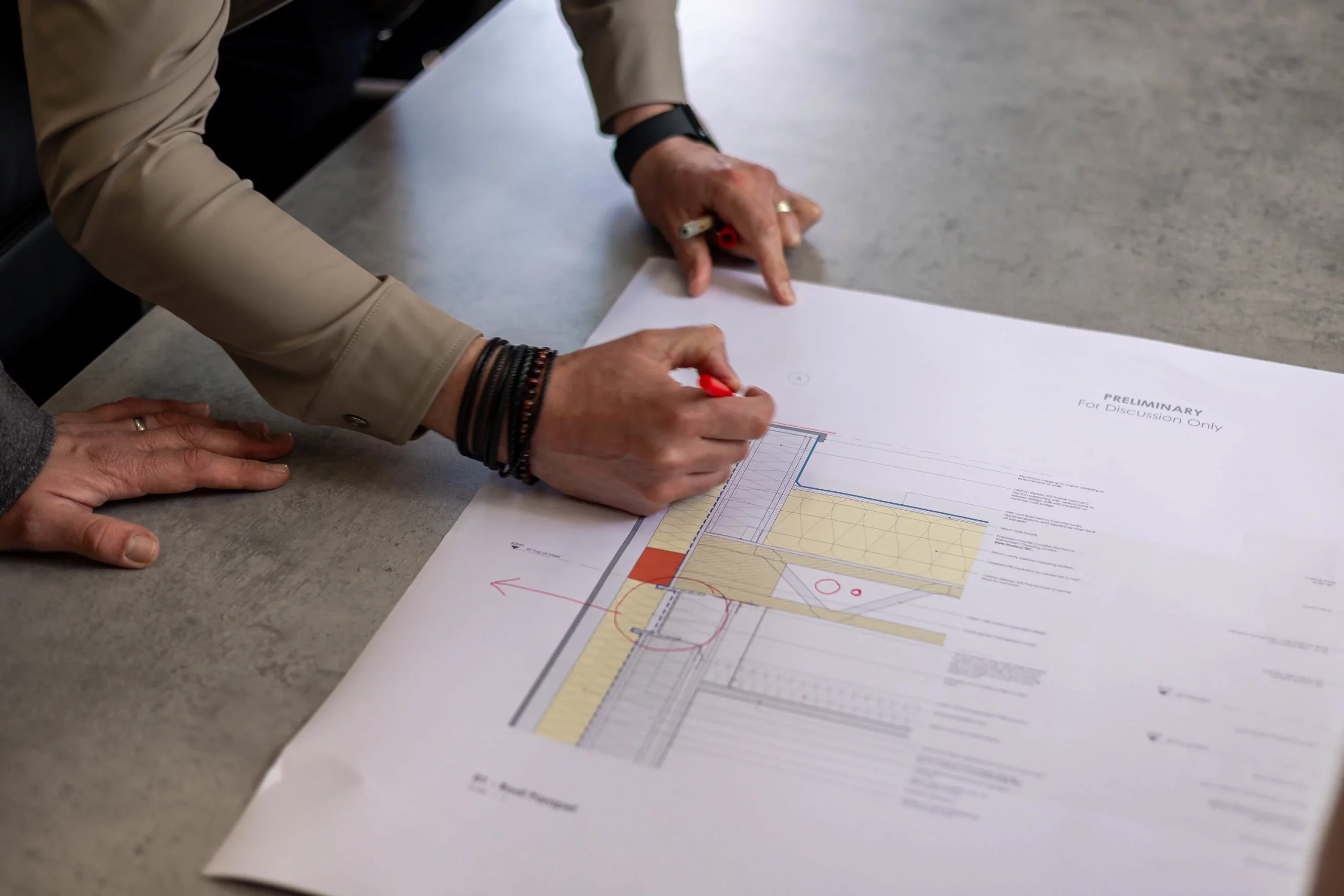 Hands pointing and marking a large architectural drawing on a table, one holding a red marker and another pointing at the plan labeled "PRELIMINARY".