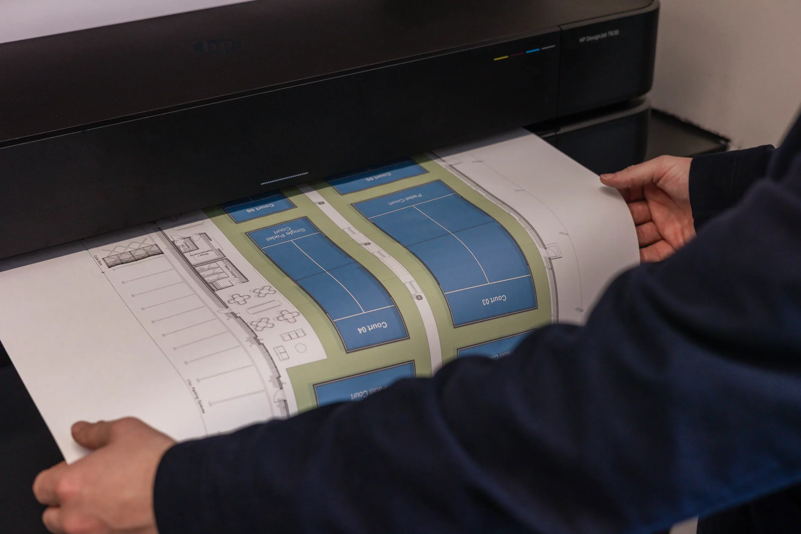 Person feeding a large colour architectural floor plan into a wide-format printer, hands holding the paper edges.