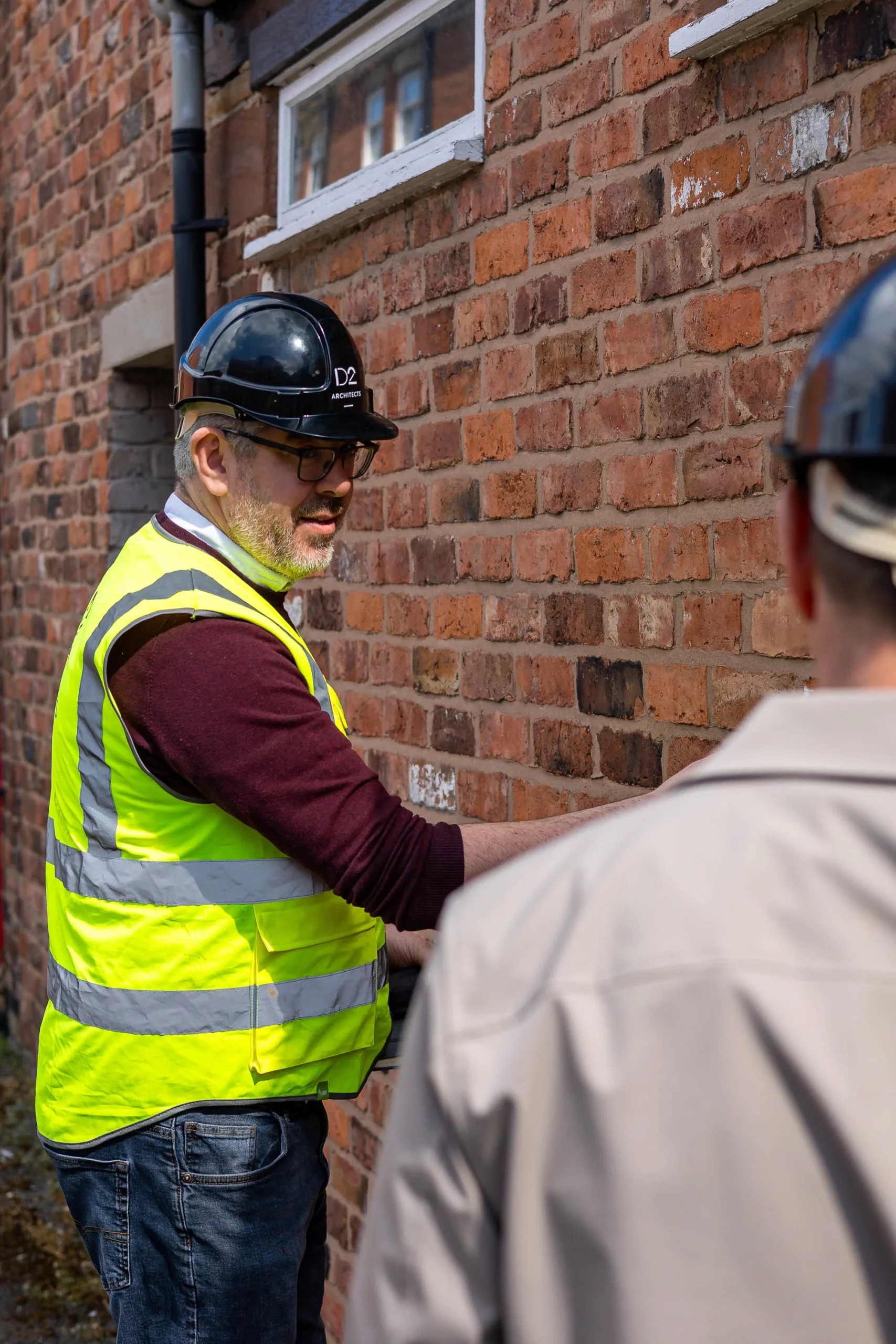 Man in a black hard hat and yellow high-visibility vest inspecting a red brick wall while talking to another worker.