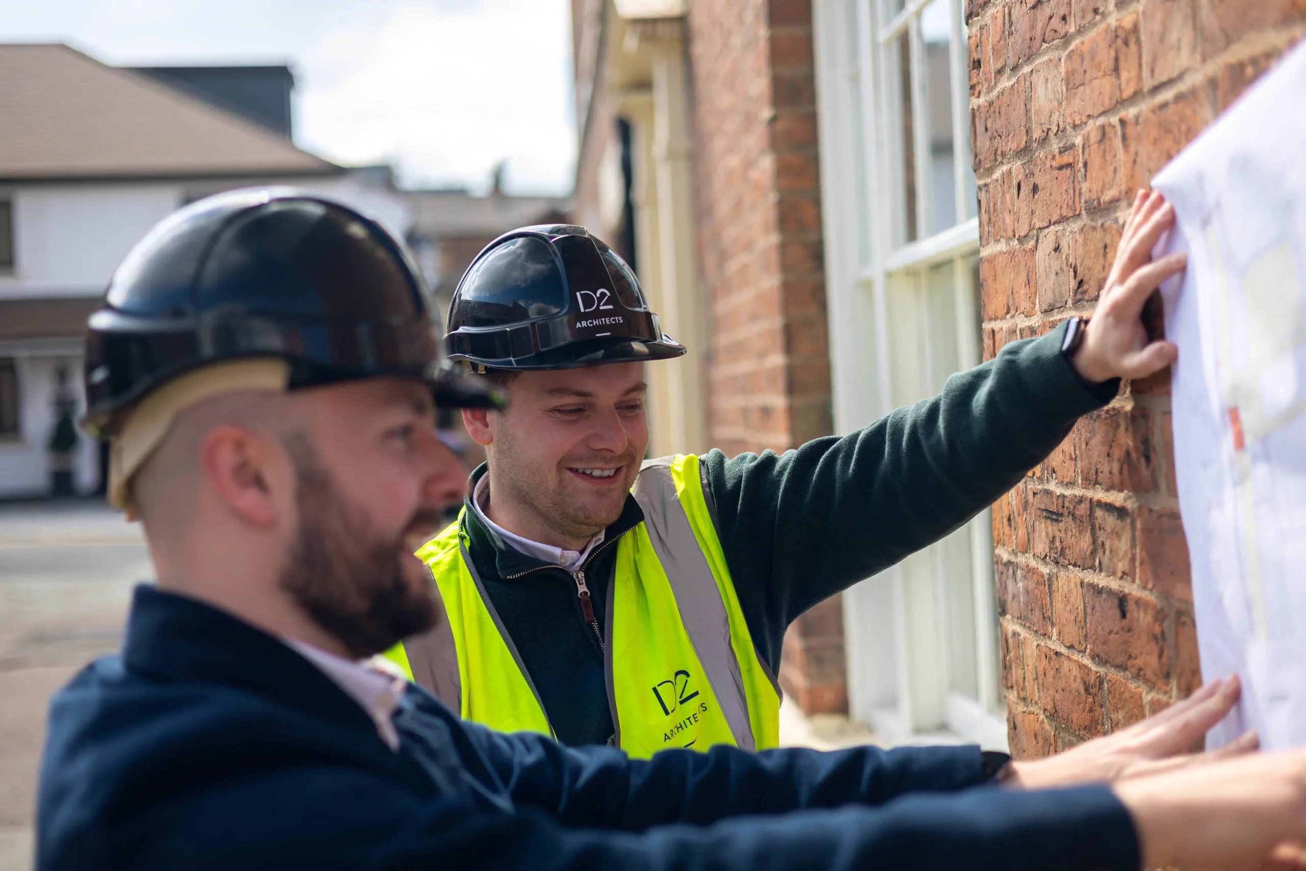 Two construction workers in hard hats and high-visibility vests examining plans taped to a brick wall, smiling