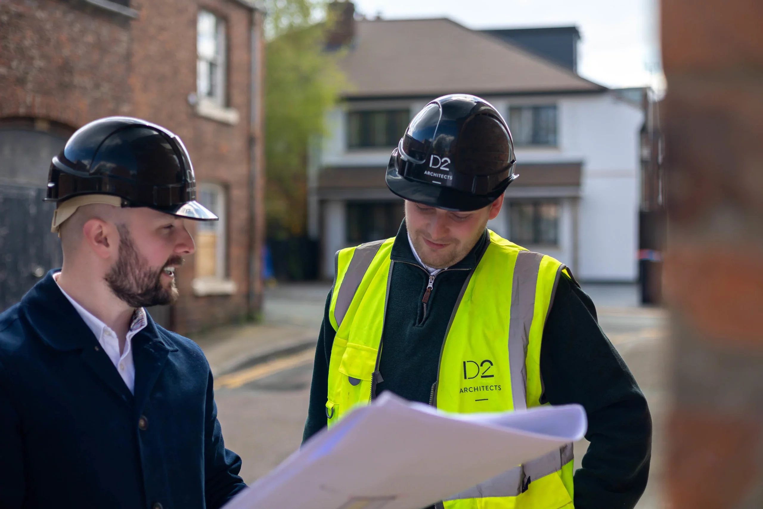 Two men in black hard hats reviewing plans outdoors, one wearing a high-visibility D2 Architects vest and looking down.