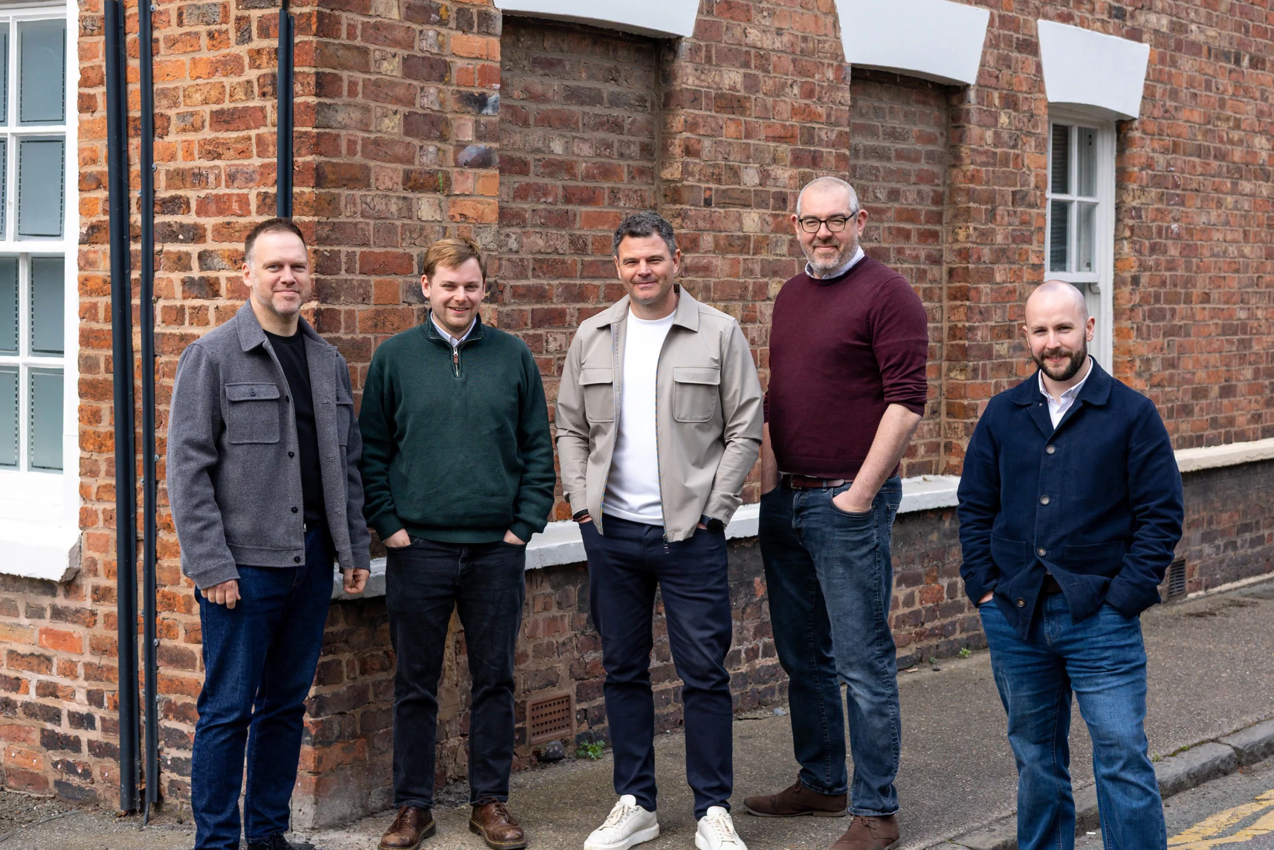 Five casually dressed men standing in front of a red-brick building, smiling and posing together.