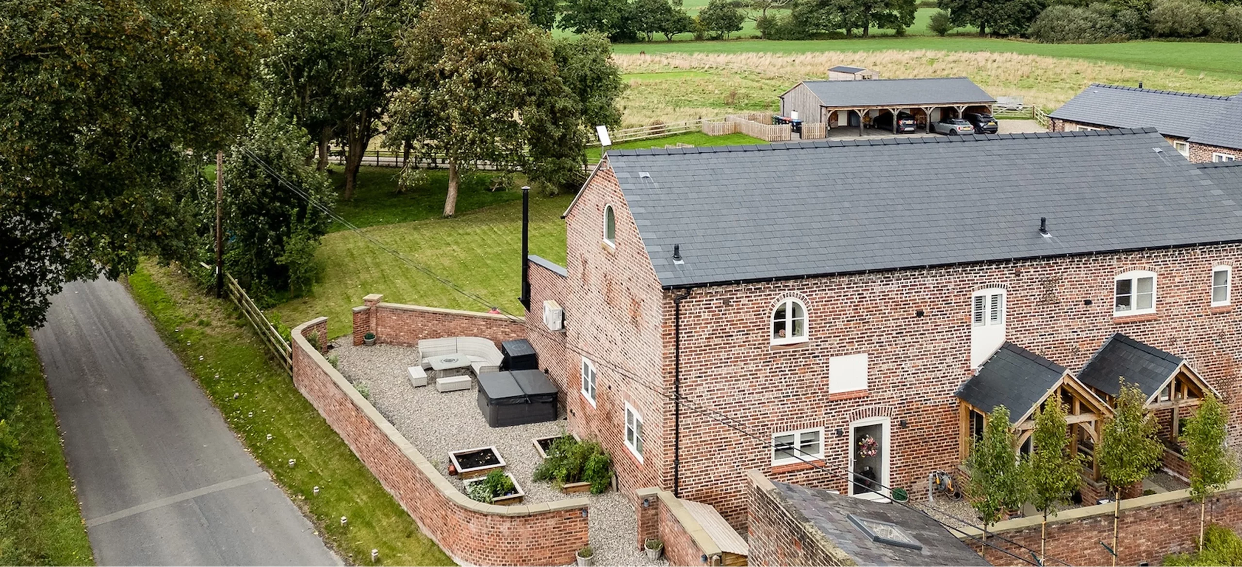 Red brick country house with slate roof, enclosed gravel courtyard with seating and planters, driveway and trees beside fields.